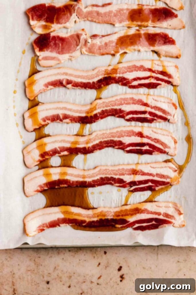 Raw bacon slices drizzled with maple syrup on a parchment-lined baking sheet, preparing for the candying process.