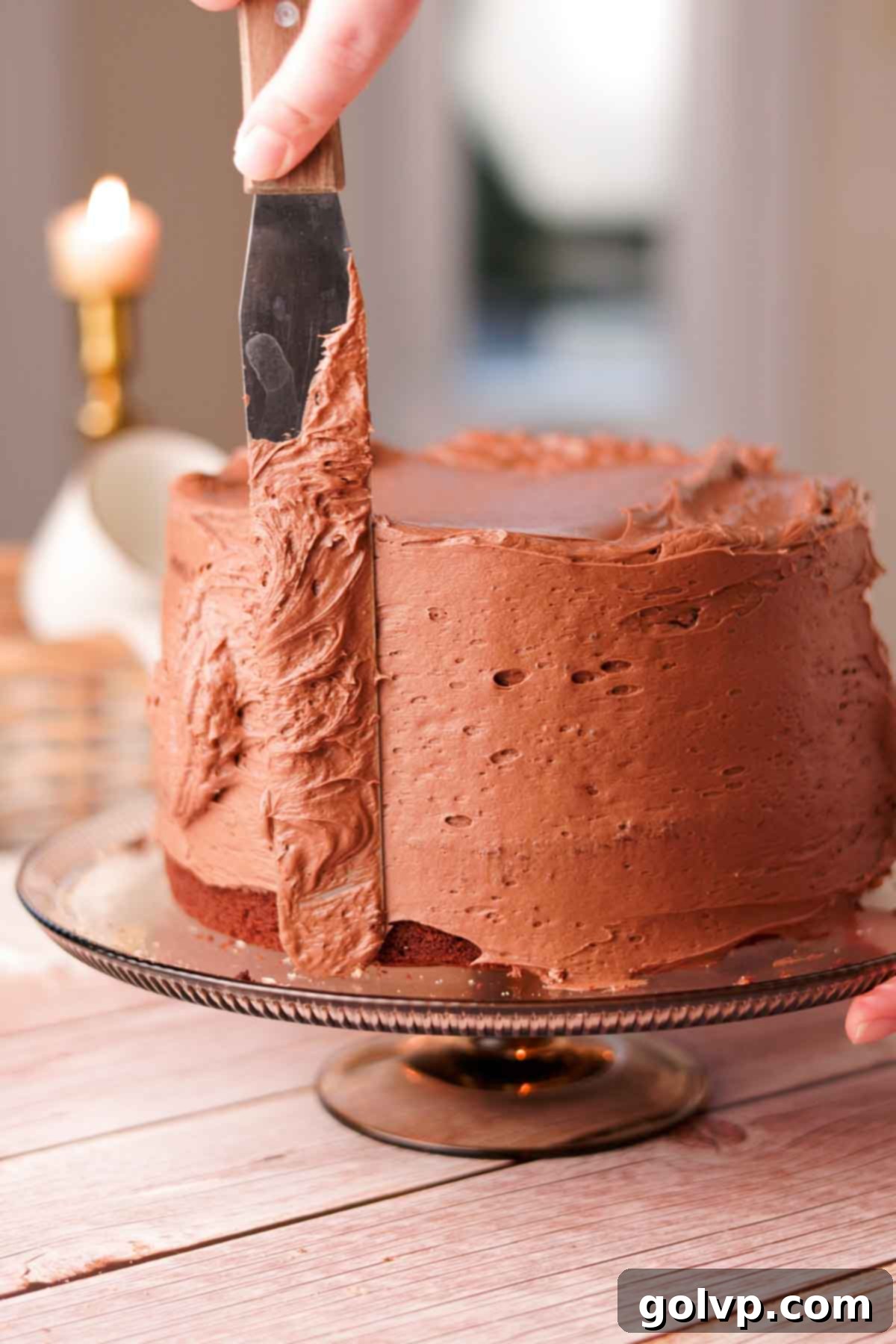 Smoothing the sides of a chocolate cake with chocolate buttercream using a long metal offset spatula on a rotating cake stand.