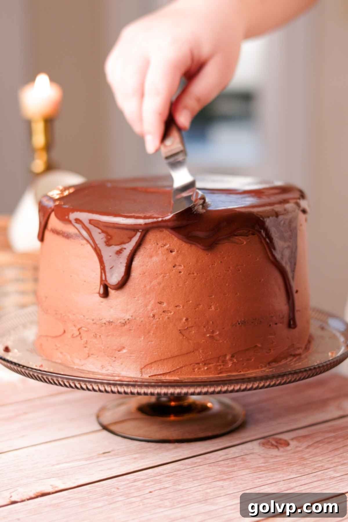 Carefully spreading an easy chocolate drip over the top edge of a chocolate cake with a small offset spatula, allowing it to naturally cascade down the sides.