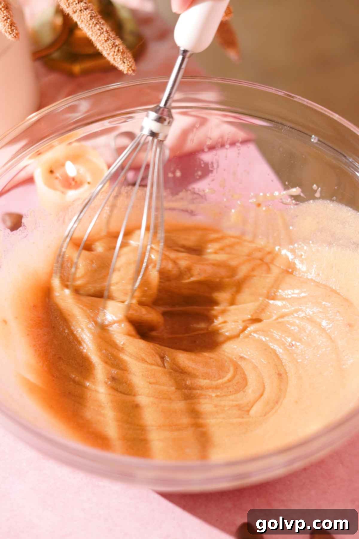 A wire whisk rapidly combines butter, sugars, and eggs in a glass bowl, creating a lightened and visibly thickened mixture.