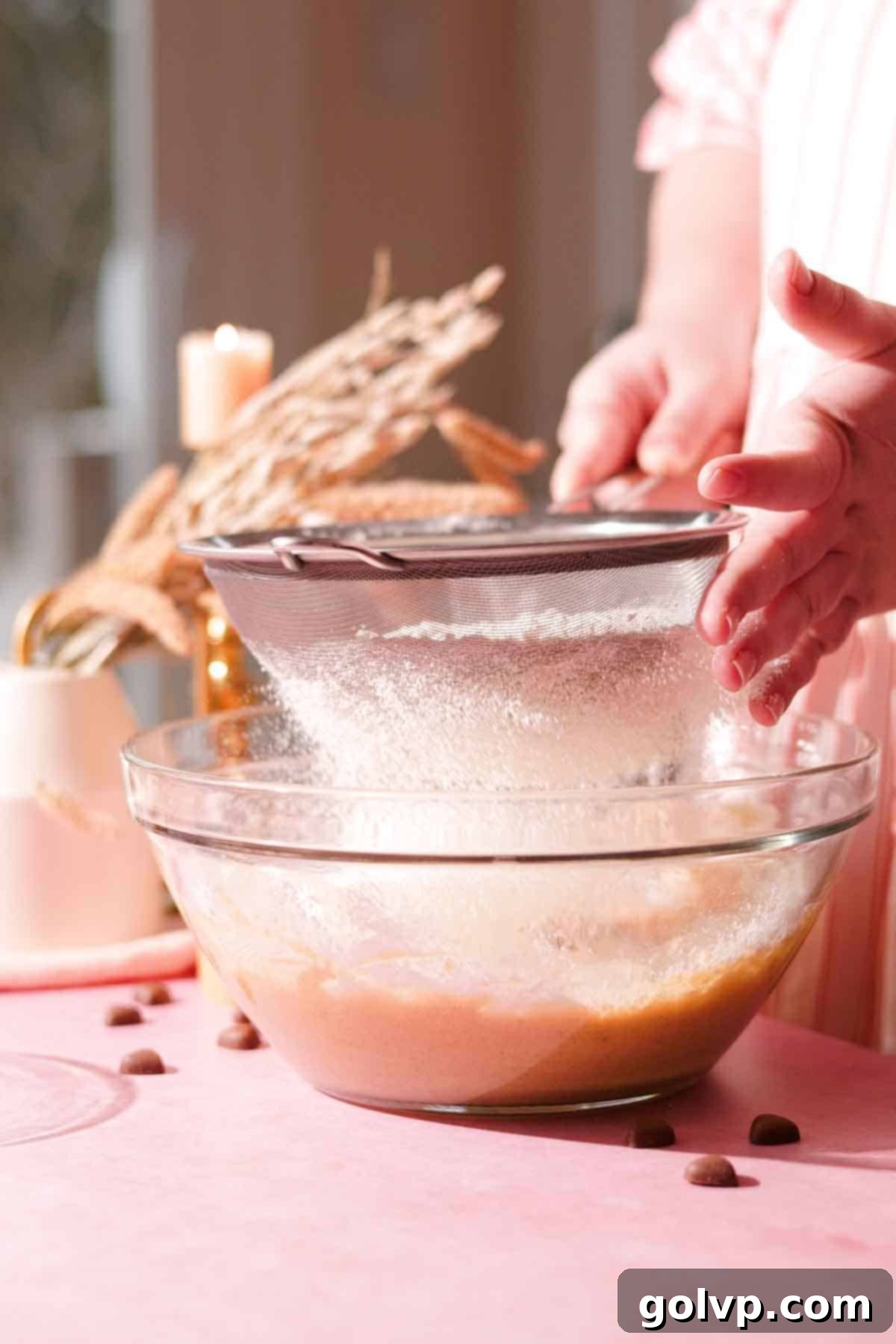 Dry ingredients, including flour and baking soda, are carefully sifted through a metal sieve directly into a glass bowl containing the wet cookie dough mixture, ensuring a smooth, lump-free batter.