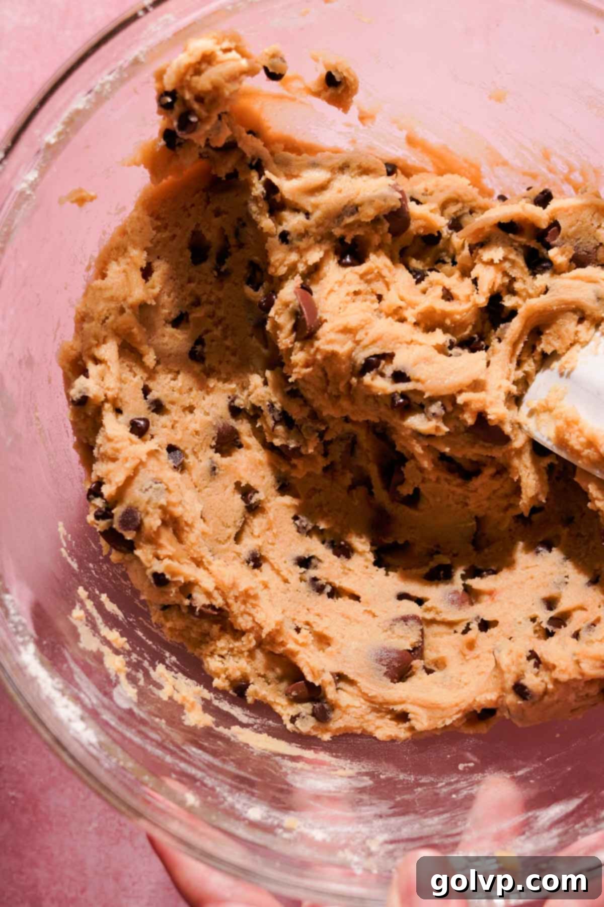 A silicone spatula gently folds chocolate chips into a rich, golden cookie dough in a glass bowl, ensuring the ingredients are just combined without overmixing.