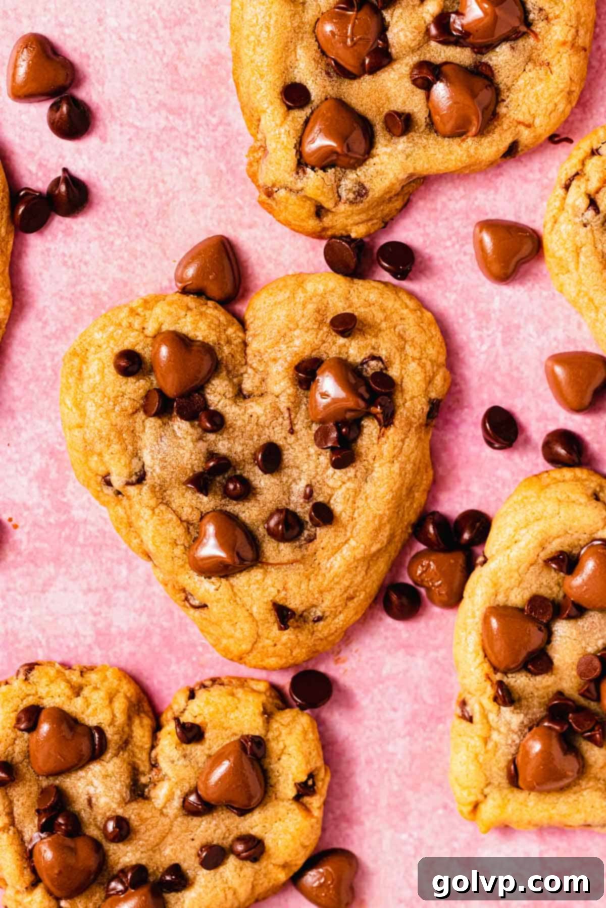Freshly baked heart shaped chocolate chip cookies arranged on a pink surface, showcasing their golden edges and soft centers, still warm and inviting.