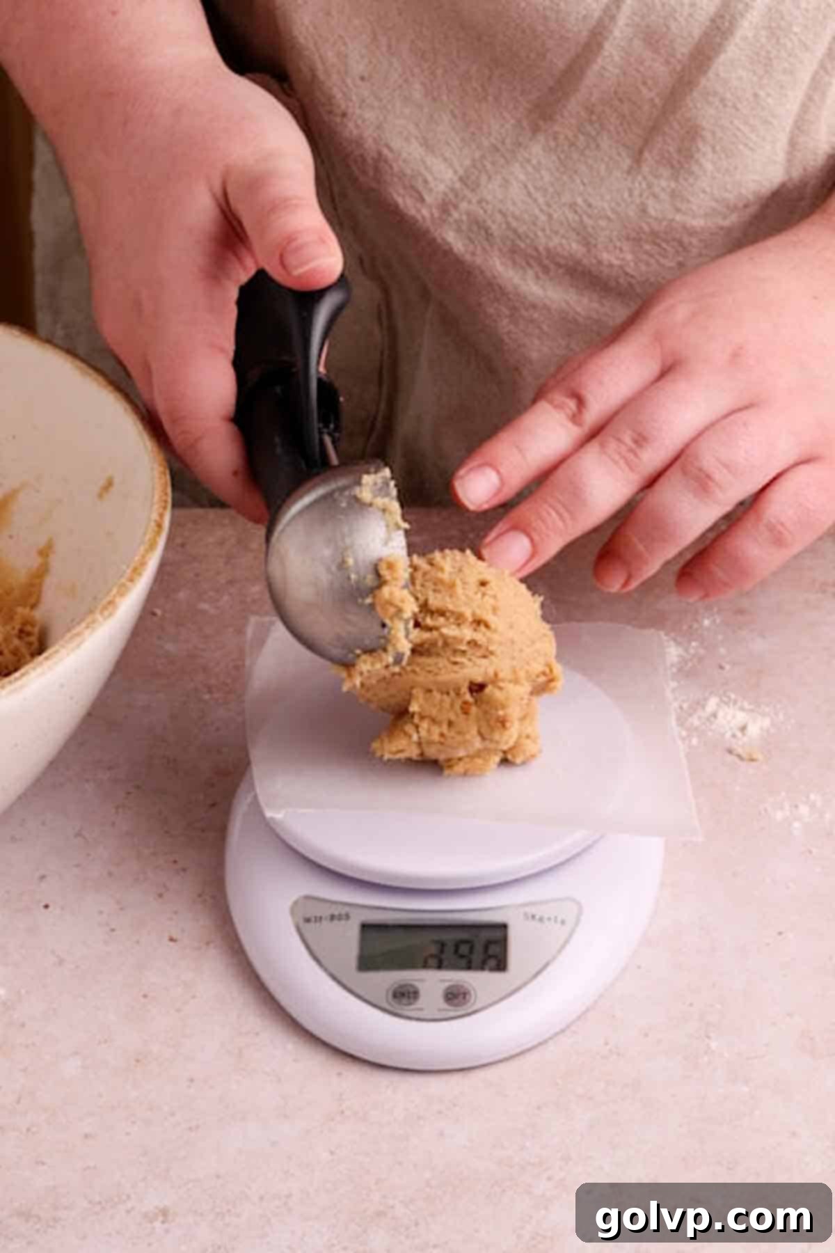 A baker carefully weighing a portion of cookie dough on a digital kitchen scale, ensuring precise measurements for consistent cookie size.