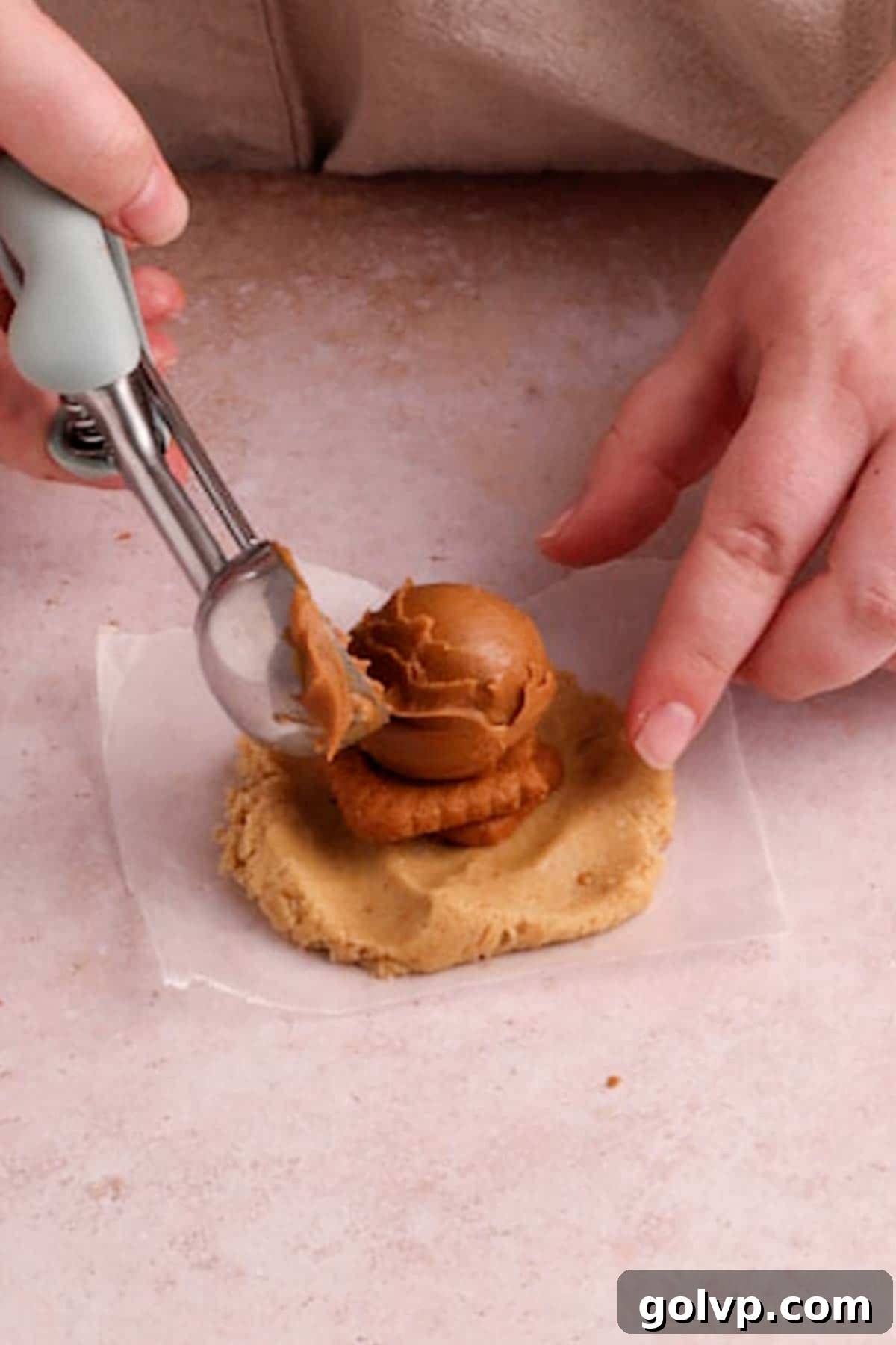 Hands carefully assembling a Biscoff stuffed cookie, placing a dollop of cookie butter and crushed Biscoff cookies onto a flattened disk of dough, ready to be enclosed by another dough disk.