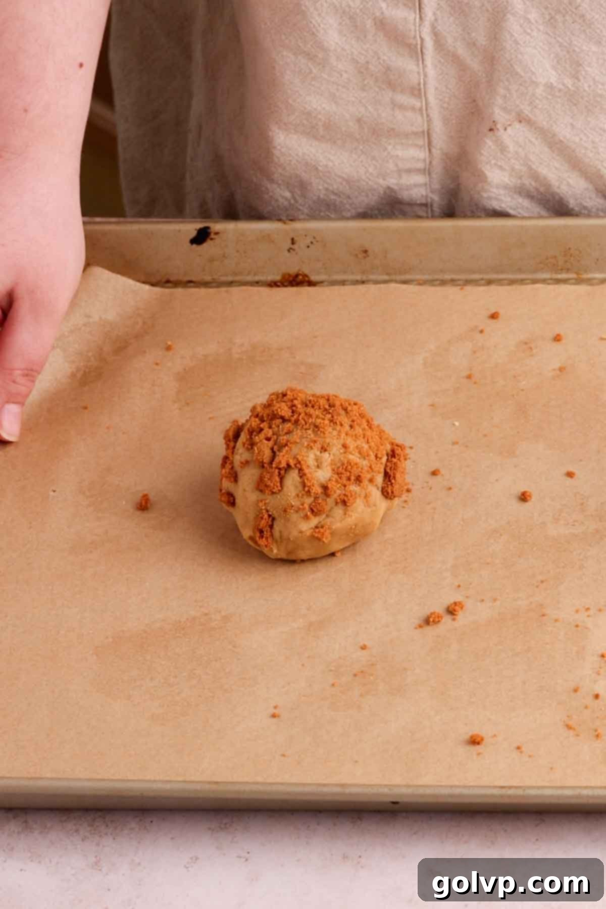 A perfectly formed, Biscoff stuffed cookie dough ball, with its top covered in crushed Biscoff crumbs, sitting on parchment paper, ready for chilling and baking.