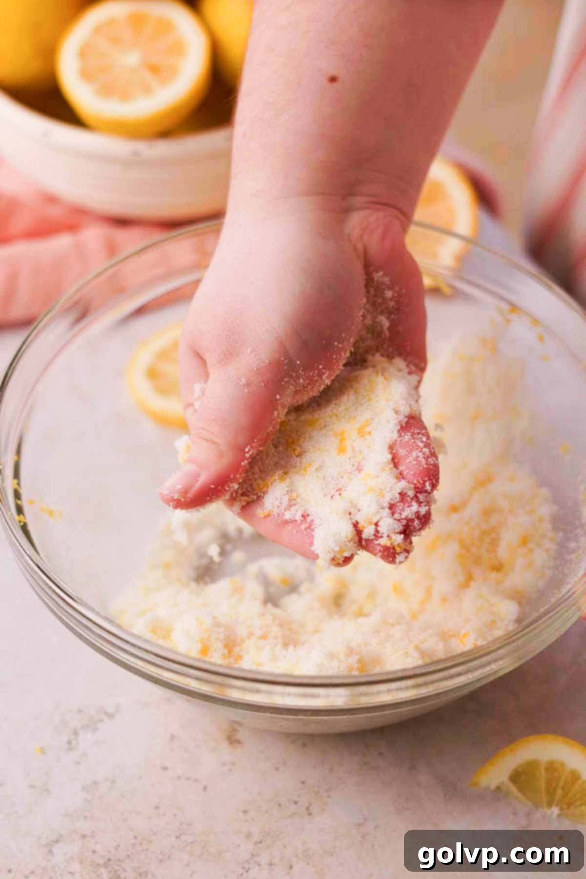 Fingers rubbing lemon zest and granulated sugar together in a large glass bowl to release aromatic oils, creating a fragrant lemon sugar.