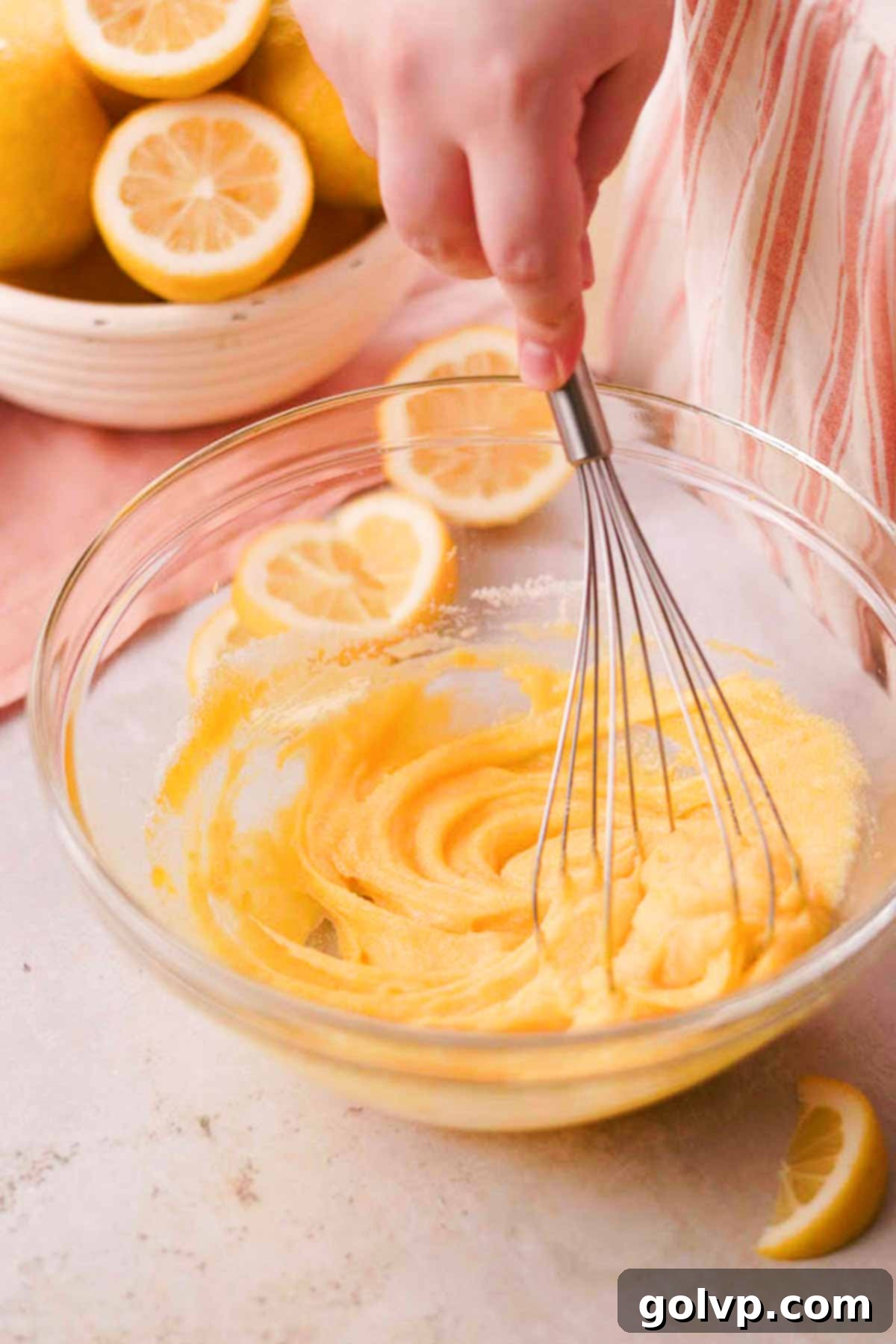 A metal whisk rapidly blending lemon-infused sugar, a whole egg, and an extra egg yolk in a glass bowl until the mixture is light and thickened.