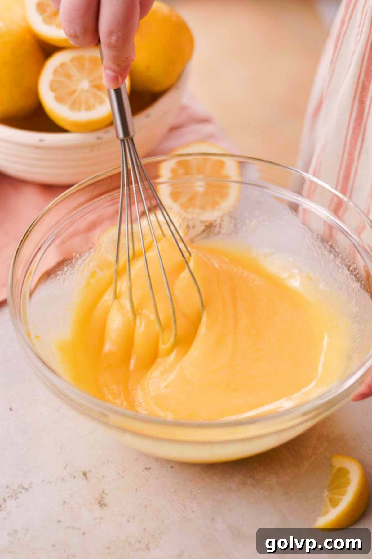 Melted and cooled butter being poured into the lemon-sugar-egg mixture in a glass bowl, then whisked by hand until smooth and emulsified.