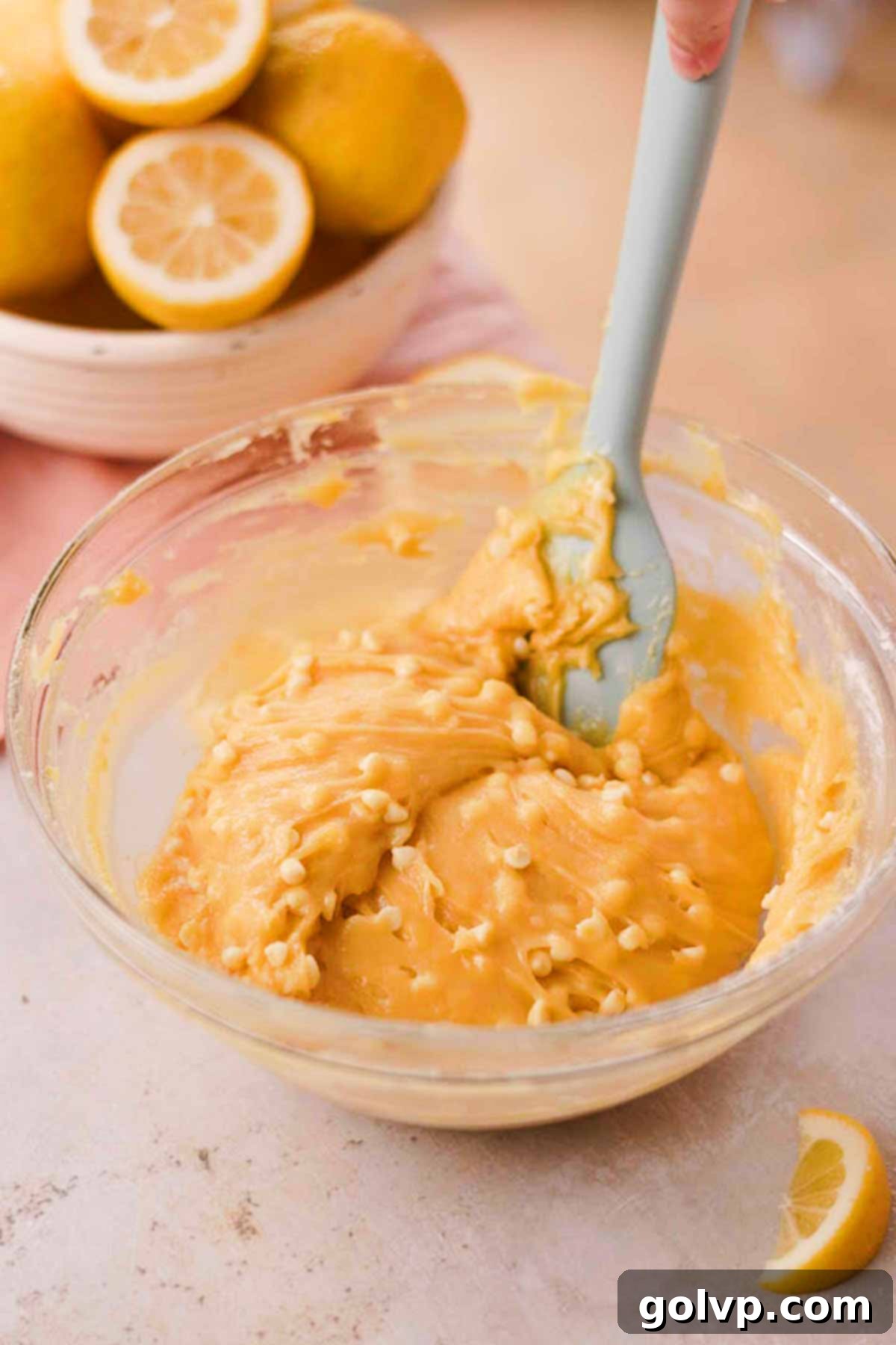 White chocolate chips being carefully folded into the lemon cookie dough with a spatula in a glass bowl.