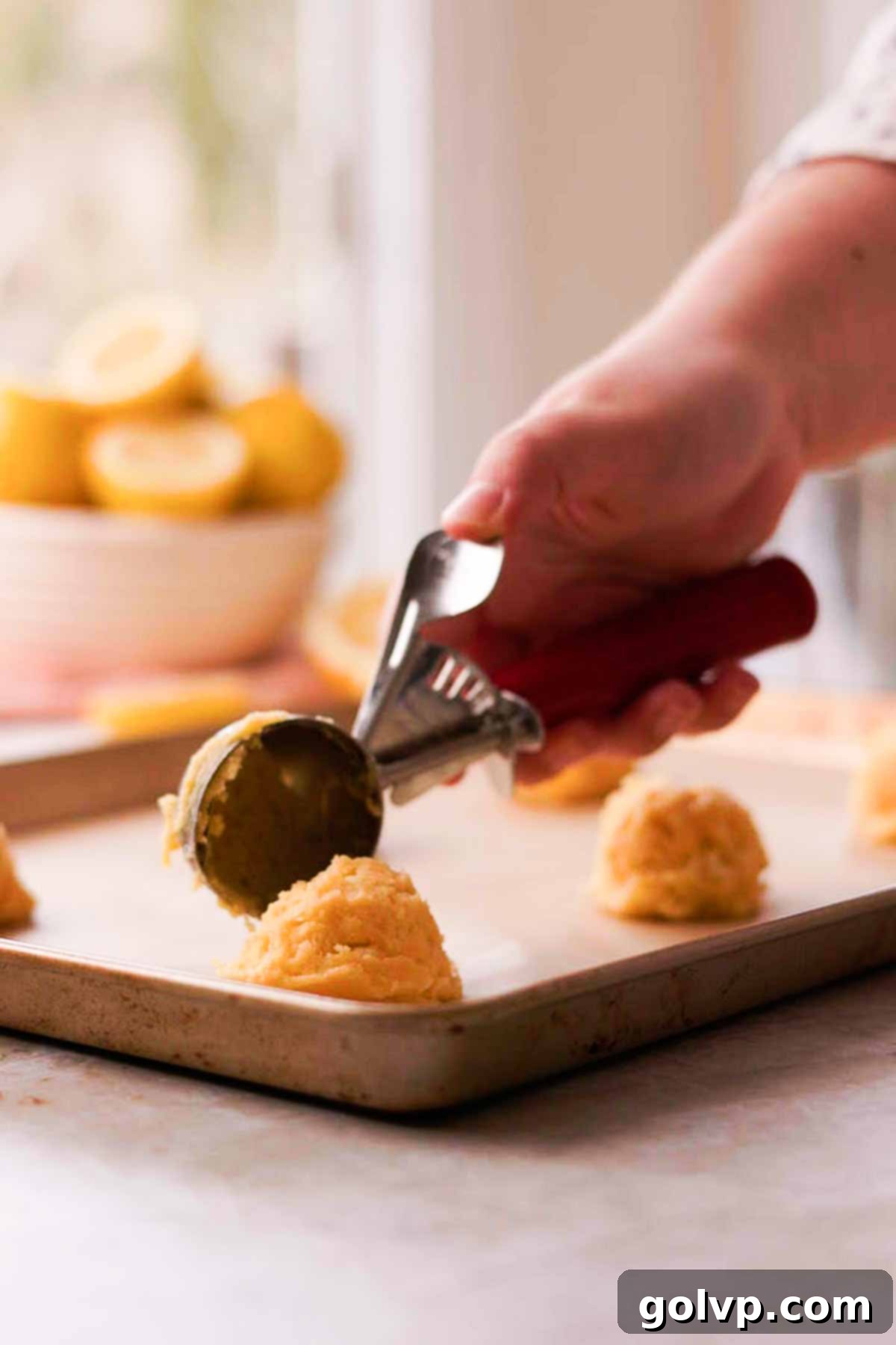 Lemon cookie dough balls being scooped and placed onto a parchment paper lined baking sheet, ready for baking.