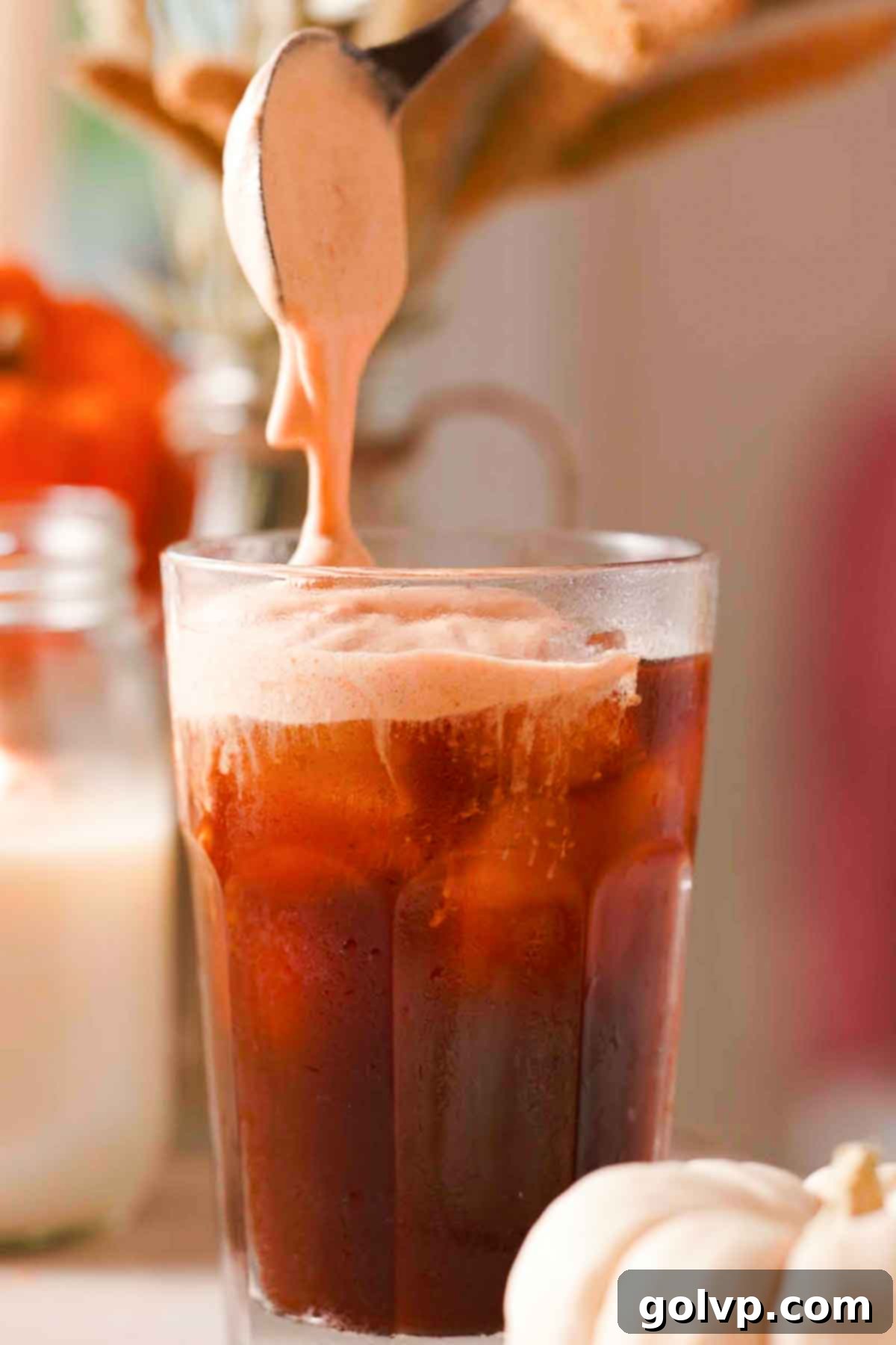Pouring freshly frothed pumpkin cold foam over an iced coffee in a tall glass, illustrating the final step of creating a delicious fall beverage.