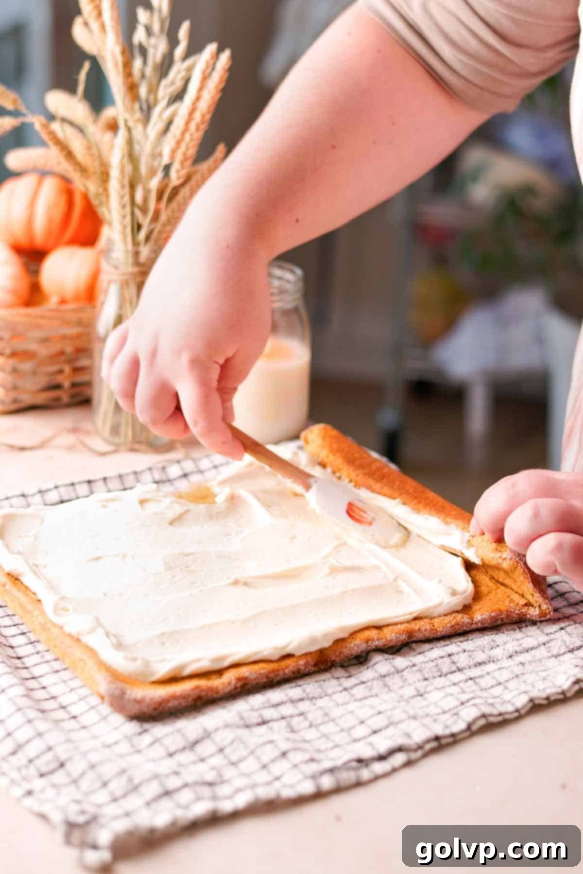 A smooth layer of creamy cheesecake filling being spread evenly onto an unrolled pumpkin sponge cake, ready for the final roll.