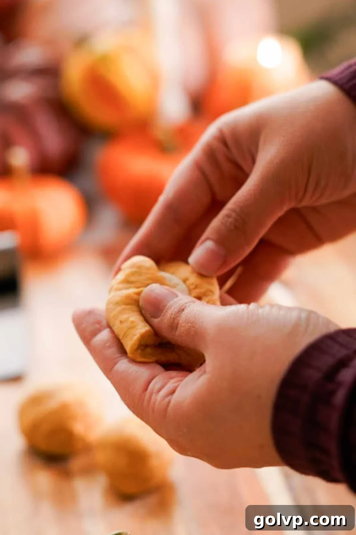 A frozen cream cheese dollop being carefully enclosed within a flattened piece of pumpkin dough, with edges pinched to seal.